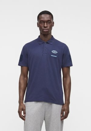 Homme portant un polo bleu marine à manches courtes avec des écussons et un pantalon gris clair, debout devant un fond blanc uni.