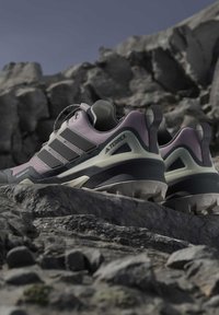 Двойка сиви и черни туристически обувки Adidas Terrex, почиващи на скалист терен с розфокусирани скални формации на заден план.