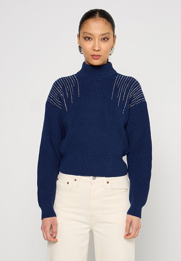 Maglione a collo alto di colore blu navy, con motivo testurizzato e dettagli lucidi sulle spalle, abbinato a pantaloni color crema.