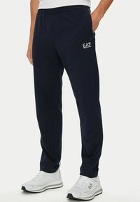 Uomo che indossa pantaloni tuta EA7 blu navy e sneakers EA7 bianche, in piedi con una mano in tasca contro uno sfondo neutro.