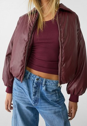 Vrouw draagt een cropped bordeauxrode jas over een bijpassend geribd tanktopje en losvallende, hooggetailleerde blauwe spijkerbroek.