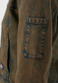 Pruun denim jakk siniste õmblustega, millel on tasku varrukal. Tekstuuritud kangas omab kulunud välimust.