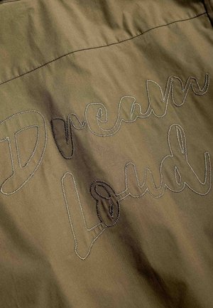 Khakinvärinen kangas, johon on kirjailtu koristeellisesti sanat "Dream Loud" kaunokirjoituksella pinnan yli.