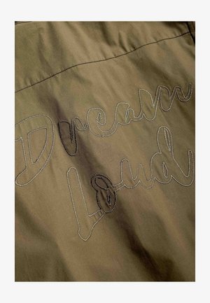 Khakistoff med broderte ord "Dream Loud" i kursiv skrift over flaten.