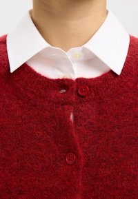 Rdeč pleten pulover z mehko teksturo, s krožnim izrezom, dvema rdečima gumboma spredaj, nošen čez belo srajco s ovratnikom.