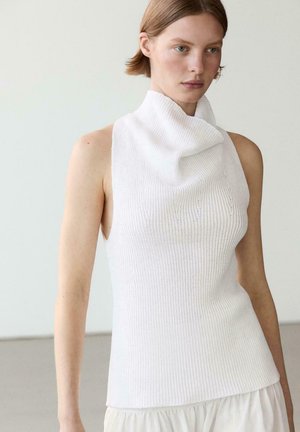 Kobieta w białym, bezrękawnikowym topie z prążkowanego dzianiny z wysokim, złożonym kołnierzem typu komin oraz w jasnej spódnicy na jednolitym tle.