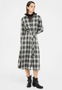 Kvinde står iført en sort og hvid ternet midikjole med bælte, med blonder ved halsudskæringen og sorte ankelstøvler.