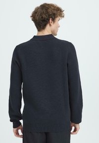 Junger Mann mit lockigem Haar, der einen schwarzen Langarmpullover trägt, von hinten vor einem schlichten hellen Hintergrund gezeigt.