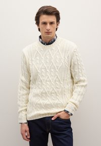 Cremfarbener Kabelstrickpullover mit Rundhalsausschnitt, strukturiertem Design und gerippten Bündchen. Über einem blauen karierten Hemd getragen; kombiniert mit dunklen Jeans.