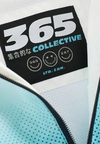 Un'etichetta in tessuto nera con la scritta "365 COLLECTIVE" in grassetto bianco e blu, circondata da icone circolari di faccine sorridenti, su un capo di colore azzurro chiaro.