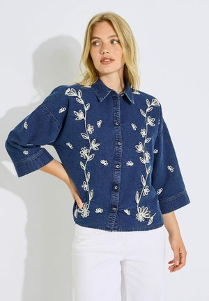 Mujer con camisa vaquera azul oscuro con bordado floral blanco y pantalones blancos, de pie con una mano en la cadera.