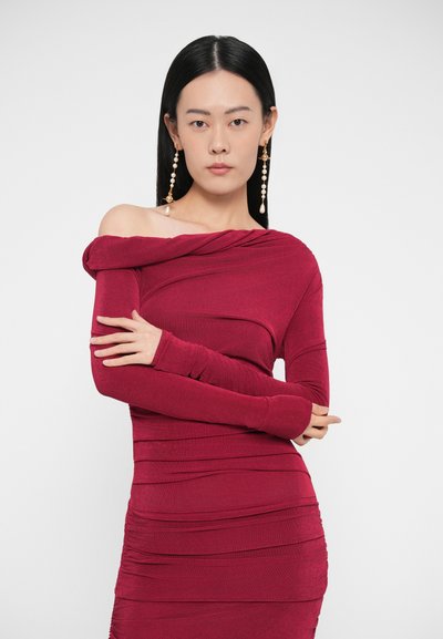 Sieviete ar gariem melniem matiem, valkājot piegulošu tumši sarkanas krāsas ārplecu kleitu ar savītiem rakstiem un garas pērļu piliens formas auskarus, rokām sakrustotām.