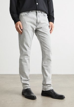 Personne portant un jean slim gris clair, des bottines en cuir noires et une chemise noire à manches longues, debout sur un sol gris.