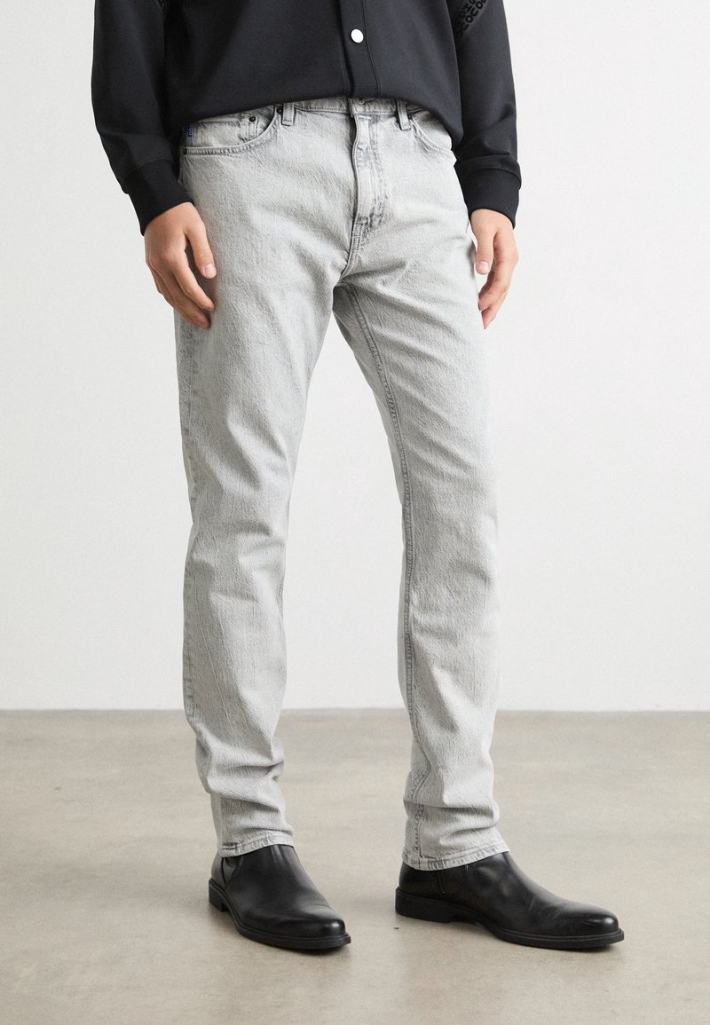 Personne portant un jean slim gris clair, des bottines en cuir noires et une chemise noire à manches longues, debout sur un sol gris.