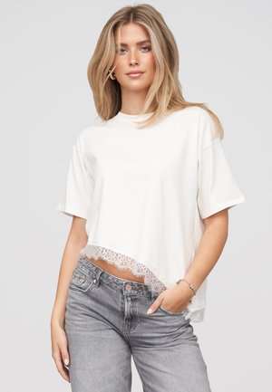 Lyshåret kvinde iført en hvid kortærmet top med blondekant og grå jeans, stående med den ene hånd i lommen mod en ensfarvet baggrund.