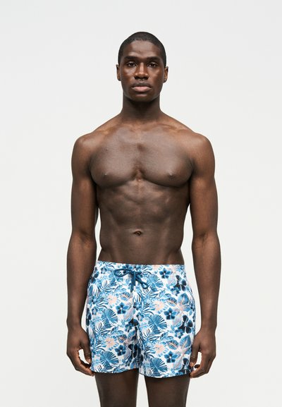 Μυώδης άνδρας που στέκεται, φορώντας μπλε και λευκά φλοράλ μαγιό με κορδόνι, μπροστά σε απλό φωτεινό φόντο.