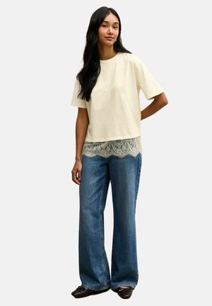 Mujer con el pelo largo y negro, vestida con una camiseta corta de manga corta color crema con bordes de encaje, vaqueros azules de pierna ancha y zapatos negros, de pie sobre un fondo liso.