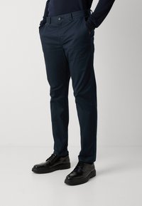Marineblauwe chino's met een slim fit, voorzien van zijzakken, een sluiting met een knop aan de voorkant en een gladde katoenen stof. Gepaard met zwarte geklede schoenen.