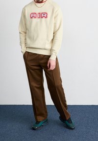 Sudadera beige de cuello redondo con el logotipo rojo "AIR" y el símbolo de Nike, combinada con pantalones marrón ajustados y deportivas verdes azules con acentos negros.