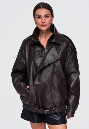 Femme portant une veste en cuir marron foncé oversize avec un col haut, une fermeture éclair, une ceinture et des poches latérales, associée à une jupe noire.