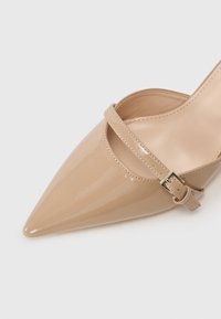 Beige patento nahkal kengät, joissa on terävä kärki, kapea nilkkaremmi metallisoljella ja kiiltävä pinta.