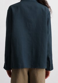 Camicia in lino di un verde scuro con una vestibilità rilassata, polsini abbottonati e una texture sottile; abbinata a pantaloni beige chiaro.
