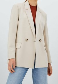 Beige blazer valmistettu sileästä kankaasta, jossa on kaksoisnapit, kaulukset ja sivutaskut. Yhdistetty vaaleanssinisiin farkkuihin.