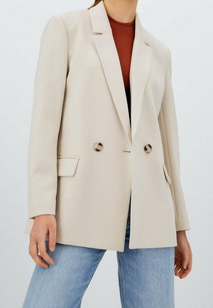 Beige blazer i släta tyg, med dubbla knappar, utskurna slag och sidofickor. Stylad med ljusblå jeans.