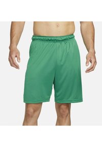 Homme portant un short de sport vert uni avec une taille élastique et des poches latérales, debout devant un fond blanc.