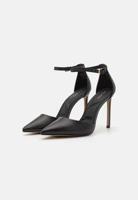 Chaussures à talons hauts noires avec un design à bout pointu, fabriquées en cuir lisse, dotées d'une bride au niveau de la cheville et d'un fin talon aiguille.