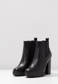 Černé kožené kotníkové boty s robustními vysokými podpatky a elastickými bočními panely, umístěné vedle sebe na bílé ploše.