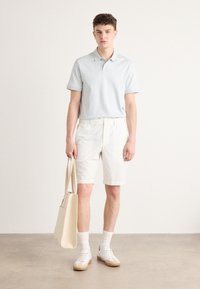 Polo light blue à manches courtes, shorts blancs et baskets blanches. Le modèle tient un sac fourre-tout beige clair, se tenant sur un fond neutre.