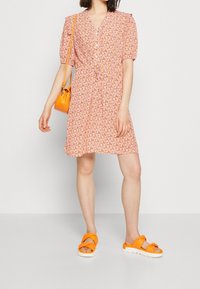 Abito rosa a fantasia floreale con maniche a sbuffo corte, scollatura a V e una cintura in vita. Abbinato a sandali arancioni chunky e una borsa coordinata.