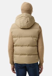 Beige puffer vest med hætte og broderet logo på ryggen, med en quiltet tekstur og parret med en beige striktrøje.
