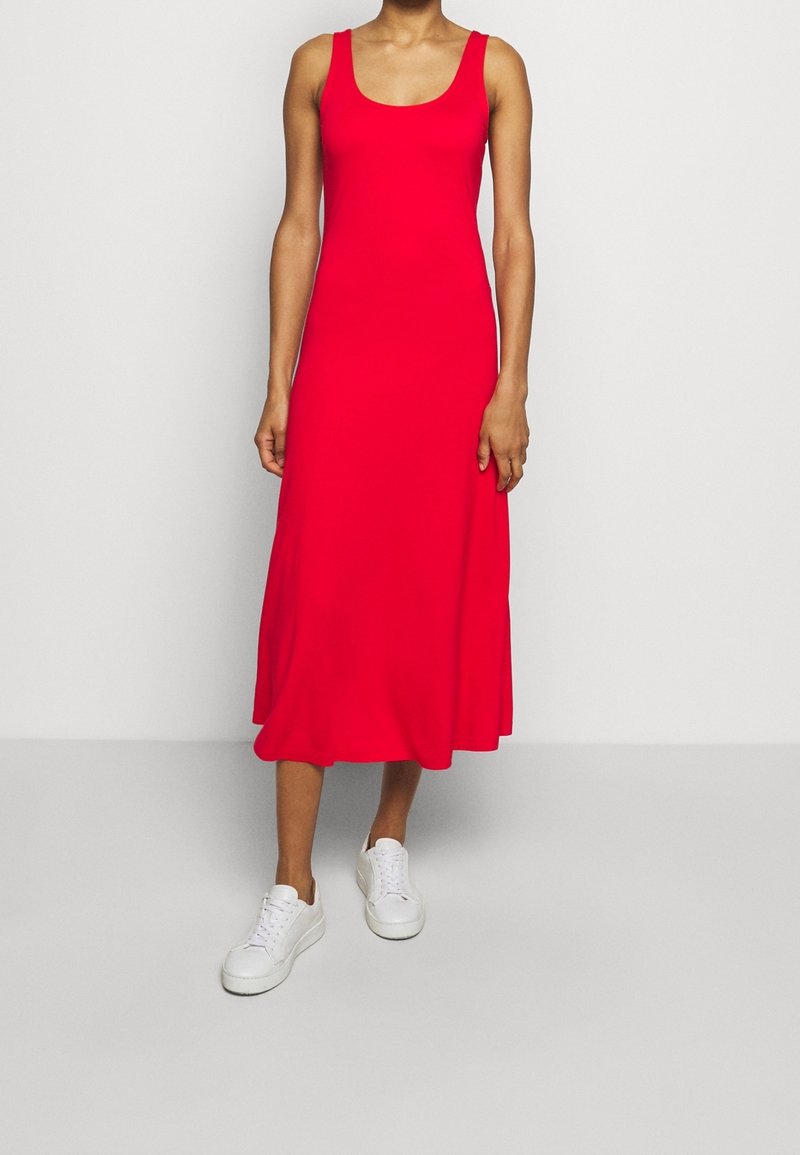 Czerwona, bezrękawnikowa sukienka midi z kopertowym dekoltem, wykonana z miękkiego materiału, o luźnym kroju, zestawiona z białymi sneakersami na jednolitym tle.