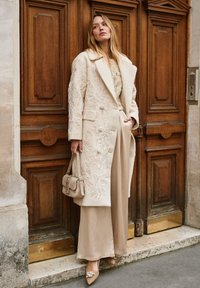 Beige embroidered coat with large lapels, paired with beige wide-leg pants. Features textured patterns and a small handbag with decorative hardware.