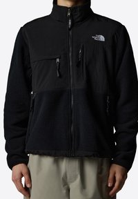 Czarna kurtka z polaru z wysokim kołnierzem, zapinana na zamek z przodu oraz wieloma kieszeniami. Posiada gładki materiał na górze i puszystą fakturę na dole.