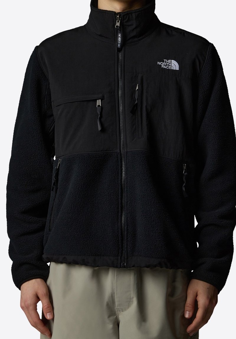 Czarna kurtka z polaru z wysokim kołnierzem, zapinana na zamek z przodu oraz wieloma kieszeniami. Posiada gładki materiał na górze i puszystą fakturę na dole.