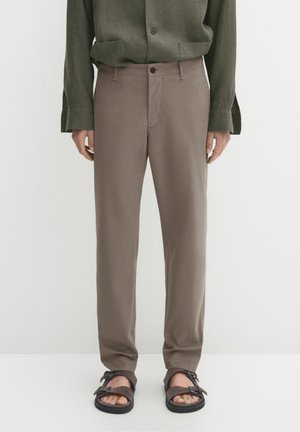 Man wearing taupe straight-fit pants paired with dark olive long-sleeve shirt and black open-toe sandals standing against white background.