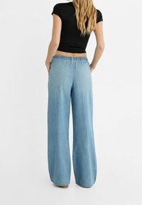 Ljusa blå vida denimbyxor med elastiskt midjeband och sidofickor, visade bakifrån mot en enkel vit bakgrund.