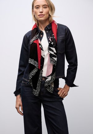 Spijkerjack over een witte blouse, gecombineerd met een kleurrijke sjaal met geometrische patronen in zwart, rood en roze tinten. Donkere jeans maken de look compleet.