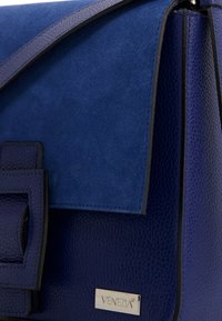 Blaues Lederhandtasche mit strukturiertem Oberflächen, Wildlederklappe, metallischem Branding und einem strukturierten Design mit einem kontrastierenden Akzent.