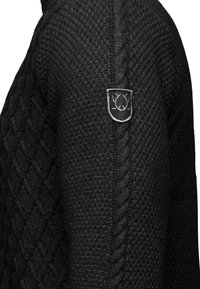 Maglione nero lavorato a maglia con texture a cavo e un logo a forma di scudo sulla spalla. Il materiale sembra spesso e caldo.