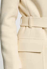 Beige wollen jas met een gestructureerde riem, textuurstof en een zijzak. De riem heeft een geribbelde uitstraling voor extra detail.