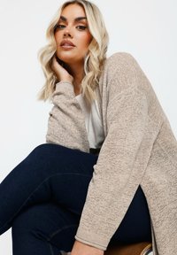 Beige stickad cardigan med långa ärmar och öppen front, matchad med mörkblå jeans. Texturen är mjuk och något ribbad.