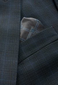 Tmavý vzorovaný materiál oblekovej bundy so subtílnym modrým kockovaným dizajnom. Svetlosivý kapesník s geometrickým vzorom je čiastočne viditeľný v vrecku.