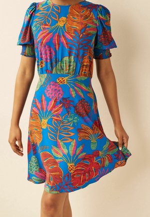Femme portant une robe bleu vif avec des motifs de feuilles tropicales orange, rouge et verte ainsi que des ananas, tenant l'ourlet d'un côté.