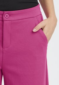 Fukszia nadrág sima textúrával, látható gombos záródással, oldalzsebbel és tiszta varrási részletekkel.
