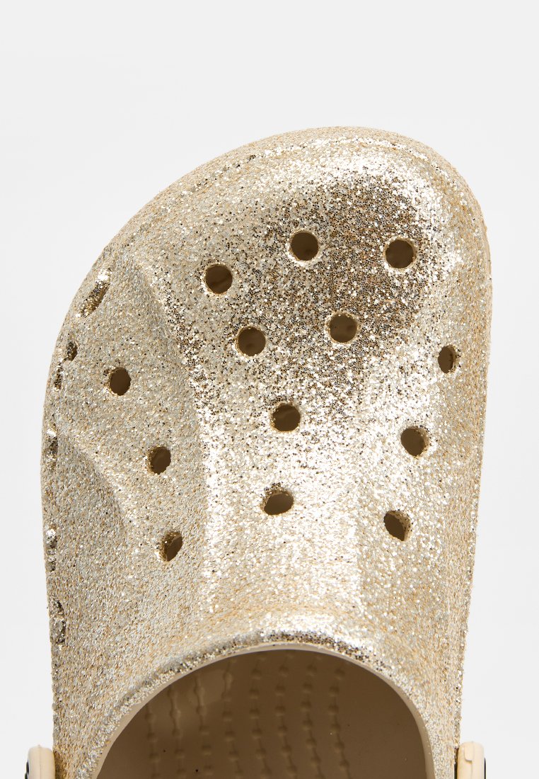 Sabots en paillettes dorées avec plusieurs trous de ventilation, bout arrondi et surface lisse et brillante ; la texture intérieure est nervurée pour le confort.