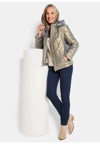 Gequiltete metallic silberne Jacke mit Kapuze, beiges Sweatshirt, dunkelblaue Hose und beige Stiefeletten, steht neben einer runden weißen Säule.